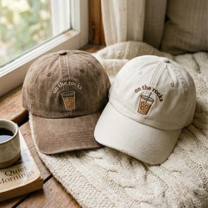 May include: Two baseball caps, one in a distressed brown and one in an off-white colour. Each cap features an embroidered graphic of an iced coffee drink with the text "on the rocks" arched above the cup in a dark brown thread colour.