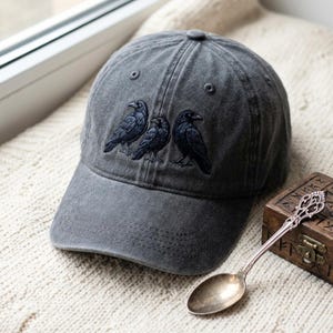 May include: A charcoal gray baseball cap featuring an embroidered design of three black ravens standing side by side on the front panel. The cap has a structured crown, a curved brim, and stitched eyelets for ventilation on the top panels.
