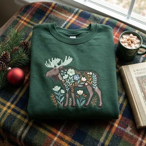 Puede incluir: Una sudadera de cuello redondo en color verde oscuro con la silueta de un alce bordada y rellena de un colorido patrón floral. El diseño del alce incluye elementos botánicos en azul claro, blanco y marrón, con flores y ramas pequeñas alrededor de las patas.