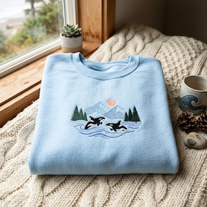 May include: A light blue crewneck sweatshirt featuring an embroidered design of two black and white orcas swimming in blue water, surrounded by green pine trees, gray mountains, and a small peach-colored sun centered on the chest area.