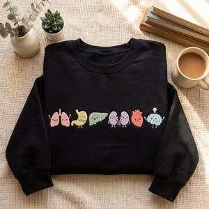 Puede incluir: Una sudadera negra de manga larga con una fila de órganos humanos de dibujos animados bordados. El diseño incluye pulmones rosas, un estómago amarillo, un hígado verde, riñones morados, un corazón rojo y un cerebro azul con una bombilla.