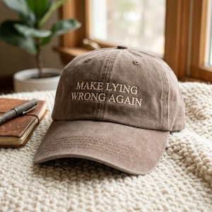 May include: A brown baseball cap with a distressed, washed-out finish. The front of the cap features the embroidered text "MAKE LYING WRONG AGAIN" in a light beige, serif font. The cap has a structured crown and a curved brim with visible stitching.