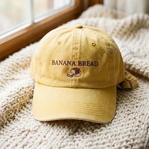 May include: A mustard yellow baseball cap featuring the embroidered text BANANA BREAD in brown thread, accompanied by a small stitched graphic of a loaf of bread and a banana. The cap has a curved brim and an adjustable metal strap at the back.