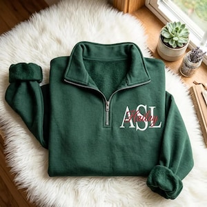 Könnte beinhalten: Ein dunkelgrünes Sweatshirt mit kurzem Reißverschluss, hohem Kragen und silberfarbenem Metallreißverschluss. Auf der linken Brustseite befinden sich die gestickten weißen Buchstaben ASL sowie der Name Hailey in roter Schreibschrift.