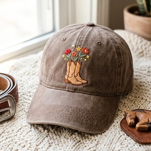 Puede incluir: Una gorra de béisbol marrón con un diseño bordado de dos botas vaqueras de color tostado llenas de flores rojas, naranjas y amarillas, acompañadas por una pequeña abeja, con visera curva y corona estructurada hecha de tela de algodón.