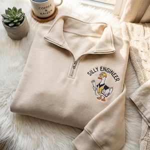 Pode incluir: Uma camisola bege com fecho de correr curto e um bordado de um ganso com capacete amarelo e colete de segurança, segurando um projeto e uma prancheta. O texto "SILLY ENGINEER" está bordado em letras pretas acima do ganso.