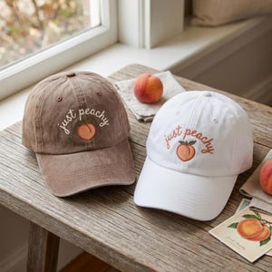 May include: A brown baseball cap and a white baseball cap, both featuring an embroidered peach graphic and the text "just peachy" in cursive lettering. Each cap has a structured crown, a curved brim, and ventilation eyelets on the top panels.
