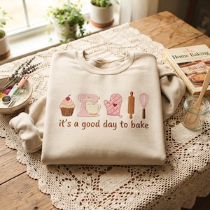 May include: A beige crewneck sweatshirt featuring embroidered kitchen icons including a cupcake, a stand mixer, an oven mitt, a rolling pin, and a whisk, with the text "it's a good day to bake" stitched in brown thread below the graphics.