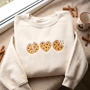 May include: A cream-colored crewneck sweatshirt featuring an embroidered design of three chocolate chip cookies: one broken in half, one heart-shaped, and one with a bite taken out of it, all rendered in shades of golden brown and dark brown.