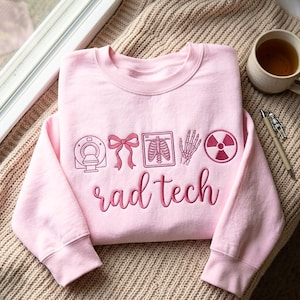 May include: A light pink crewneck sweatshirt featuring dark pink embroidery. The design includes a CT scanner, a bow, a rib cage X-ray, a hand skeleton, and a radiation symbol, with the text "rad tech" written in a cursive script below.