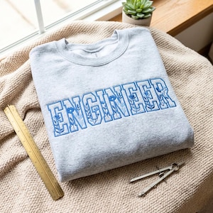 May include: A light grey crewneck sweatshirt featuring the word ENGINEER embroidered in blue block letters. The letters contain a floral pattern. A gold metal ruler and a metal drafting compass are positioned next to the sweatshirt.