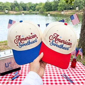 Puede incluir: Dos gorras de béisbol con coronas color crema y texto bordado que dice American Sweetheart en letra roja y azul. Una gorra tiene una visera azul sólida y la otra una visera roja sólida. Ambas incluyen pequeños detalles de estrellas.
