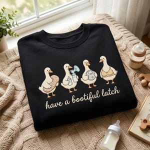 May include: A black crewneck sweatshirt featuring a graphic of four cartoon ducks. The first holds a nursing pillow, the second a breast pump, the third a pump motor, and the fourth wears a nursing bra. The text reads have a bootiful latch.