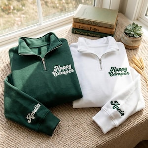 May include: A dark green quarter-zip sweatshirt with white embroidered text reading Happy Camper and the name Amelia on the sleeve, alongside a white quarter-zip sweatshirt with dark green embroidered text reading Happy Camper and Annie on the sleeve.