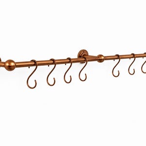 May include: A bronze-coloured metal pot rack with eight curved hooks. The rack is mounted on a white wall and features decorative end pieces and a central sphere. The hooks are evenly spaced along the horizontal bar.