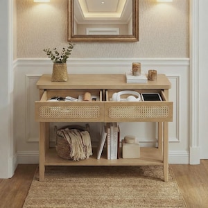 Peut inclure: Une console en bois clair avec deux tiroirs à façades en canne tressée. Les tiroirs sont ouverts, révélant des objets à l'intérieur. Un panier tressé, des livres et des objets décoratifs sont sur l'étagère inférieure. Un miroir est accroché au-dessus.