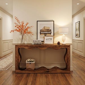 May include: A rustic wooden console table with a wavy design, holding a framed piano print, a lamp, and a small basket of flowers. A vase with orange branches and a stack of books are also on the table. The room has hardwood floors and a neutral color palette.