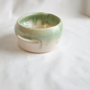 May include: A small, handmade ceramic bowl with a rounded shape. The interior and upper rim are glazed in a glossy, light green color, with the glaze dripping down the sides onto the off-white base. The bowl sits on a white surface.