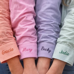 Op de afbeelding: Vier shirts met lange mouwen in perzik, roze, lavendel en mintgroen, met de namen "Gracie", "Mary", "Becky" en "Jodie" geborduurd op de manchetten. De shirts worden gecombineerd met een blauwe jeans.