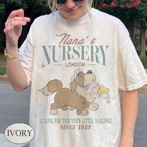 May include: An ivory t-shirt featuring a graphic of a brown dog wearing a white bonnet and carrying a baby in a pink outfit. The text reads Nana's NURSERY LONDON, CARING FOR YOU YOUR LITTLE DARLINGS SINCE 1953. The design uses brown, green, and pink colours.