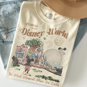 Może przedstawiać: Kremowa koszulka z grafiką Disney World na Florydzie. Projekt zawiera Myszka Miki, Hollywood Tower, zamek Kopciuszka i Spaceship Earth. Tekst na koszulce brzmi "Disney World" i "The Most Magical Place On Earth."