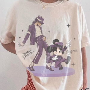 Pode incluir: Uma t-shirt de cor marfim com uma impress&atilde;o gr&aacute;fica de Michael Jackson e Mickey Mouse, ambos vestidos com fatos roxos e chap&eacute;us fedora. O design inclui o texto Michael Jackson e Mickey Mouse com pequenas estrelas. Decote redondo.