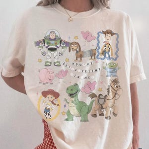 Pu&ograve; includere: T-shirt color avorio con personaggi dei cartoni animati di un popolare film d'animazione. Il design include Buzz Lightyear, Woody, Jessie, Rex, Hamm, Slinky Dog, Bullseye e i piccoli omini verdi. Viene visualizzata anche la frase "YOU'VE GOT A FRIEND IN ME".