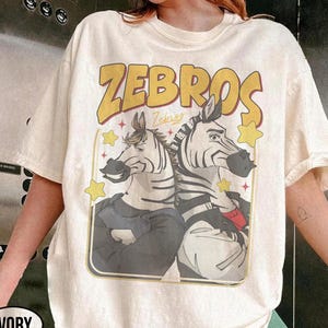 Op de afbeelding: Ivoor kleurig t-shirt met een afbeelding van twee zebra's met mensachtige trekken, omlijst door een gele rand. Het woord "ZEBROS" staat in een gele retro-lettertype boven de zebra's gedrukt. Het shirt heeft korte mouwen.