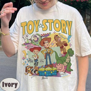 May include: Ivory-coloured t-shirt with the words "TOY STORY" in yellow, featuring cartoon characters including Buzz Lightyear and Woody. The shirt has a crew neck and short sleeves.