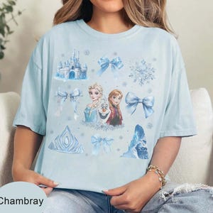 May include: Pale blue t-shirt with a design featuring characters and elements from a popular animated film. The design includes a castle, bows, snowflakes, and the text "Chambray".