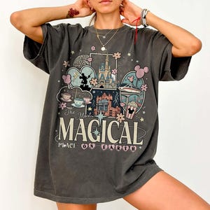 Puede incluir: Camiseta gris oscuro con un gr&aacute;fico colorido con el texto "The Most Magical Place on Earth". El dise&ntilde;o incluye ilustraciones de un castillo, tazas de t&eacute;, un monorra&iacute;l y palomitas de ma&iacute;z.