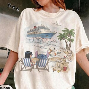 May include: Ivory-colored t-shirt featuring a beach scene with a cruise ship, palm trees, and cartoon characters. Two characters are in striped beach chairs. A sandcastle, starfish, and life preservers are also visible. The word "IVORY" is printed on the bottom left.