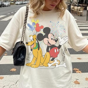 May include: Cream-colored t-shirt featuring Mickey Mouse and Pluto cartoon characters with colorful balloons and fireworks. The words "Mickey Mouse" and "Pluto" are printed on the shirt. The shirt is a casual, oversized fit.