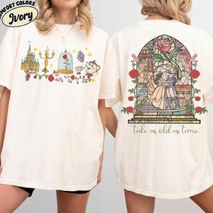 Puede incluir: Camiseta de color marfil con una ilustración colorida de un castillo, una rosa y otros elementos de cuento de hadas. La parte trasera de la camiseta muestra un diseño de vidriera con una pareja bailando, con el texto "tale as old as time" debajo.