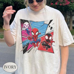 K&ouml;nnte beinhalten: Elfenbeinfarbenes T-Shirt mit einer farbenfrohen Comic-Grafik von Spider-Man-Charakteren. Das Design zeigt Spider-Man, Spider-Gwen und Miles Morales in Action-Posen, vor dem Hintergrund blauer, rosa und schwarzer Panels. Das Wort "IVORY" ist unten aufgedruckt.