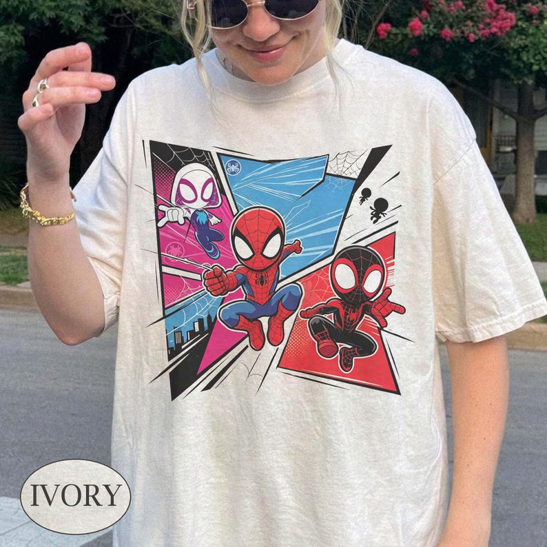 K&ouml;nnte beinhalten: Elfenbeinfarbenes T-Shirt mit einer farbenfrohen Comic-Grafik von Spider-Man-Charakteren. Das Design zeigt Spider-Man, Spider-Gwen und Miles Morales in Action-Posen, vor dem Hintergrund blauer, rosa und schwarzer Panels. Das Wort "IVORY" ist unten aufgedruckt.