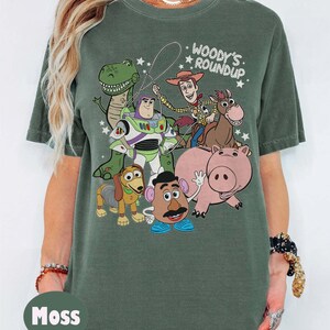 May include: Moss green t-shirt featuring a graphic of characters from the animated film "Toy Story." The design includes Woody, Buzz Lightyear, Rex, Hamm, Mr. Potato Head, Slinky Dog, and Bullseye. The text "Woody's Roundup" is above the characters.