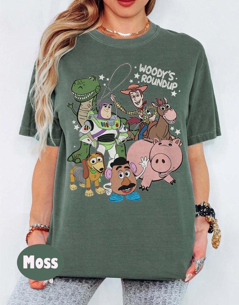 May include: Moss green t-shirt featuring a graphic of characters from the animated film "Toy Story." The design includes Woody, Buzz Lightyear, Rex, Hamm, Mr. Potato Head, Slinky Dog, and Bullseye. The text "Woody's Roundup" is above the characters.