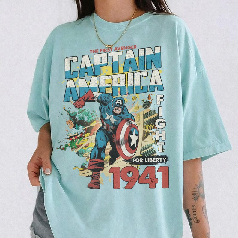 Puede incluir: Una camiseta azul claro con un estampado gr&aacute;fico de Capit&aacute;n Am&eacute;rica en una pose din&aacute;mica con un escudo. El texto dice THE FIRST AVENGER CAPTAIN AMERICA FIGHT FOR LIBERTY 1941. El dise&ntilde;o utiliza colores rojo, blanco, azul y amarillo.