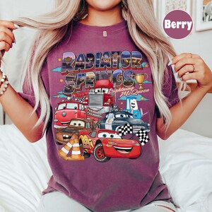 May include: Berry-coloured t-shirt with a vibrant graphic of Cars movie characters, including Lightning McQueen and Mater. The words "Radiator Springs" are above the characters, with a trophy and chequered flags.