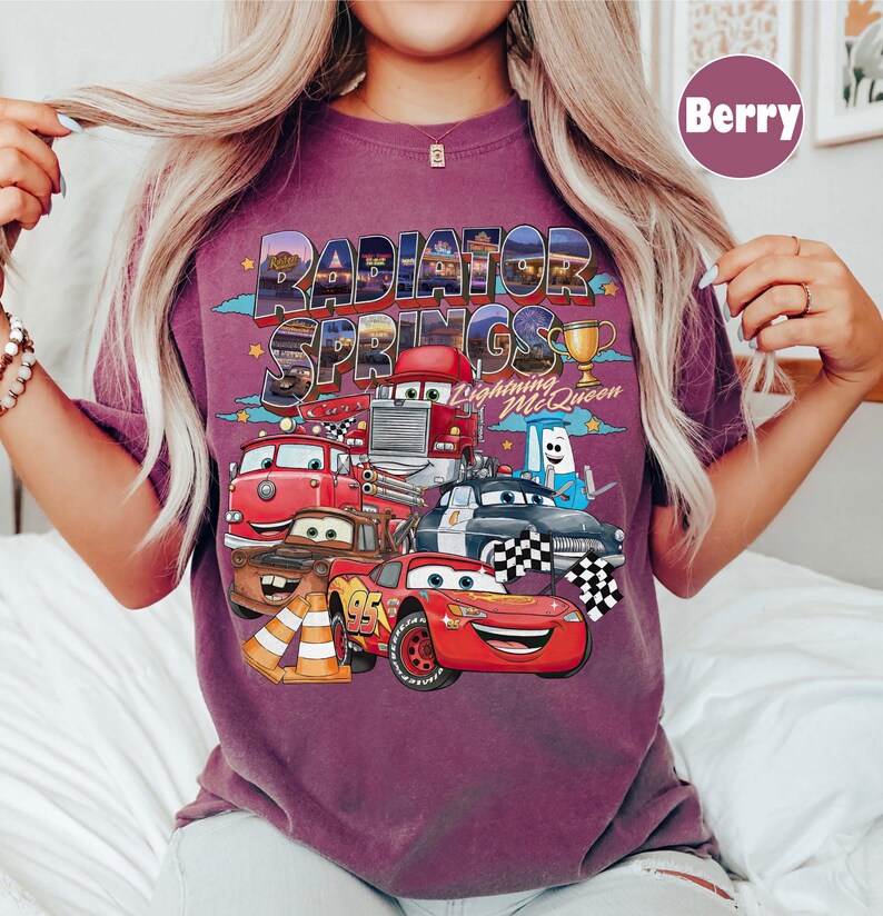 May include: Berry-coloured t-shirt with a vibrant graphic of Cars movie characters, including Lightning McQueen and Mater. The words "Radiator Springs" are above the characters, with a trophy and chequered flags.