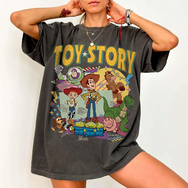 May include: Dark grey oversized t-shirt with the words "TOY STORY" in yellow and a colourful graphic of characters from the animated film. The design includes Woody, Buzz Lightyear, Jessie, and other characters. The shirt has a vintage, worn look.