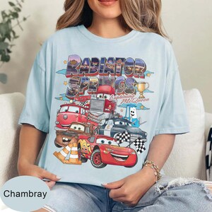 May include: Pale blue t-shirt with a vibrant graphic of Cars movie characters, including Lightning McQueen. The words "RADIATOR SPRINGS" are printed above the characters. A gold trophy is also visible.