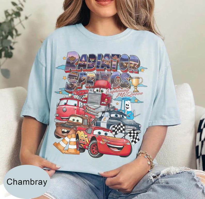 May include: Pale blue t-shirt with a vibrant graphic of Cars movie characters, including Lightning McQueen. The words "RADIATOR SPRINGS" are printed above the characters. A gold trophy is also visible.