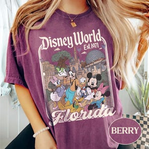 May include: Berry-colored t-shirt featuring a Disney World graphic. The graphic includes the text "Disney World Est. 1971" and a colorful illustration of Disney characters and landmarks. The word "Florida" is also displayed.
