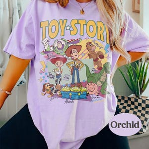 May include: A lilac t-shirt with a yellow "TOY STORY" logo and a cartoon graphic of characters from the film. The graphic includes Woody, Buzz Lightyear, Jessie, and other characters. The shirt has short sleeves.
