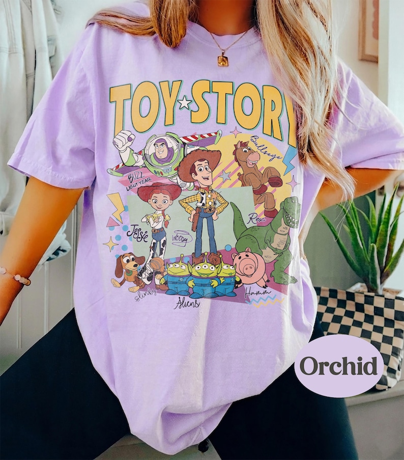 May include: A lilac t-shirt with a yellow "TOY STORY" logo and a cartoon graphic of characters from the film. The graphic includes Woody, Buzz Lightyear, Jessie, and other characters. The shirt has short sleeves.
