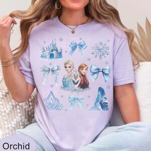 May include: Lilac t-shirt with a design from the film "Frozen", featuring Elsa, Anna, and Olaf, alongside blue bows, snowflakes, and a castle. The word "Orchid" is printed at the bottom of the shirt.