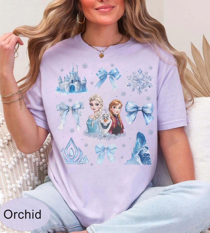 May include: Lilac t-shirt with a design from the film "Frozen", featuring Elsa, Anna, and Olaf, alongside blue bows, snowflakes, and a castle. The word "Orchid" is printed at the bottom of the shirt.