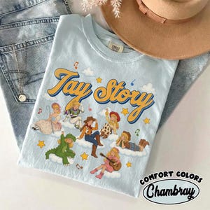 May include: A light blue t-shirt featuring the text Tay Story in yellow and blue script. The graphic includes multiple illustrated figures dressed in various costumes, including a dinosaur, a cowgirl, and a space ranger, all positioned on white clouds with yellow stars and musical notes.
